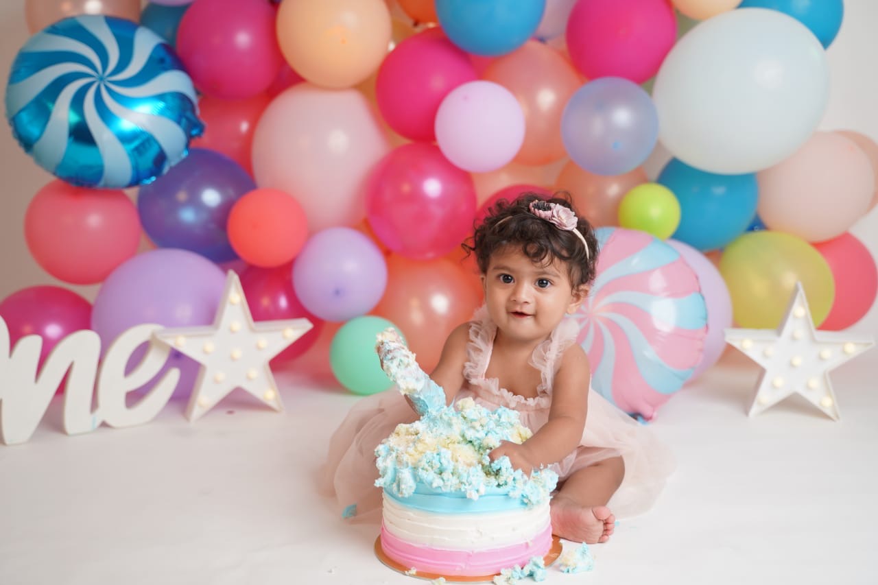 Baby Yanvi with a messy pastel cake in front of a wall of colourful balloons